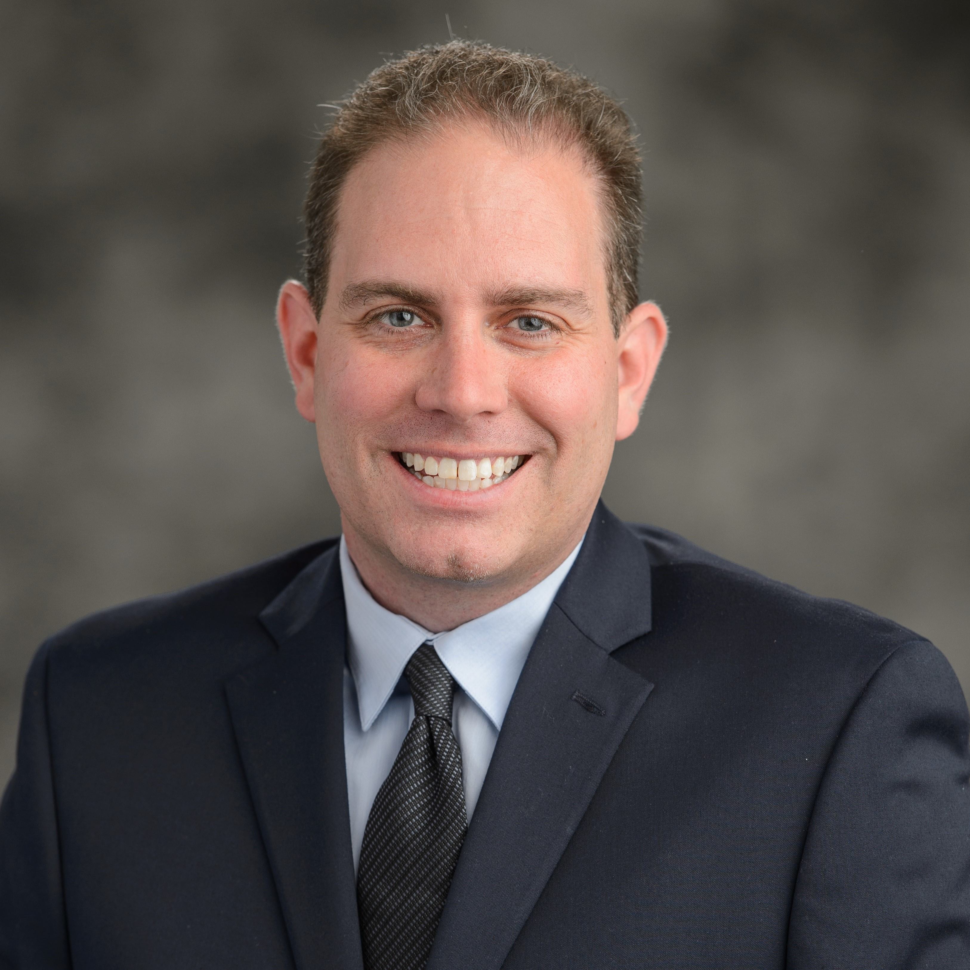 Andrew Schwarz is wearing a blue suit. He is against a dark grey background.