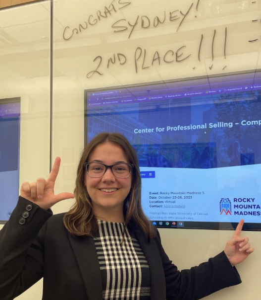 Woman with glasses and black jacket holds up an "L" with fingers and points to a computer screen.